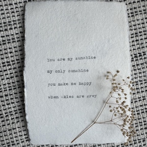 Pu&ograve; includere: Un foglio di carta ruvida bianco sporco con le parole "You are my sunshine, my only sunshine, you make me happy, when skies are grey" stampate in nero. Un rametto di fiori secchi &egrave; appoggiato sulla carta, su uno sfondo a motivi.