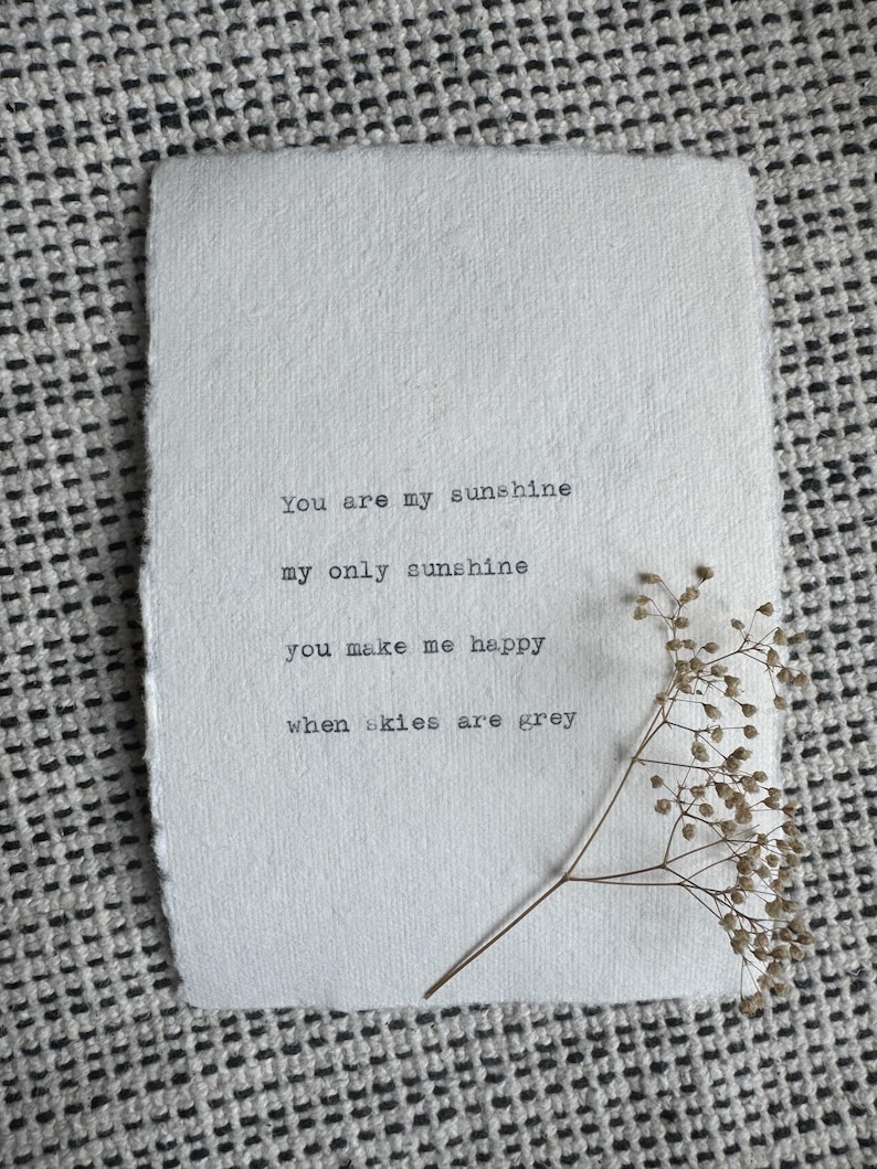 Pu&ograve; includere: Un foglio di carta ruvida bianco sporco con le parole "You are my sunshine, my only sunshine, you make me happy, when skies are grey" stampate in nero. Un rametto di fiori secchi &egrave; appoggiato sulla carta, su uno sfondo a motivi.