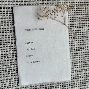 Könnte beinhalten: Ein strukturiertes weißes Papier mit den Wörtern "YOUR TEXT HERE", "quotes", "lyrics", "poems" und "love notes" in Schwarz gedruckt. Ein Zweig getrockneter Blumen liegt auf dem Papier. Das Papier liegt auf einer schwarz-weißen Webfläche.