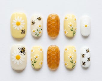 Puede incluir: Un juego de diez uñas postizas con diversos diseños que incluyen margaritas amarillas y blancas, pequeñas abejas negras y amarillas, y un patrón de panal de color ámbar texturizado sobre un fondo amarillo claro y blanco.