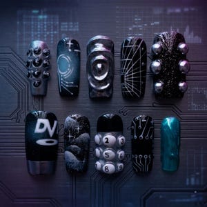 May include: A collection of ten artificial nails with various designs. The nails feature a range of colors including silver, black, and teal. Designs include geometric patterns, celestial motifs, and the letters "DV". Some nails have 3D embellishments.