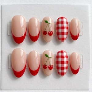 May include: A set of ten almond-shaped press-on nails featuring a nude base. The designs include red French tips, red cherry illustrations with green stems, and a red and white gingham checkered pattern on a glossy, smooth surface finish.