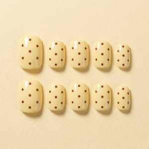 May include: A set of ten press-on nails in a light cream color, each featuring a repeating pattern of small, dark brown polka dots. The nails are arranged in two rows of five, displaying a variety of sizes with a smooth, glossy finish.
