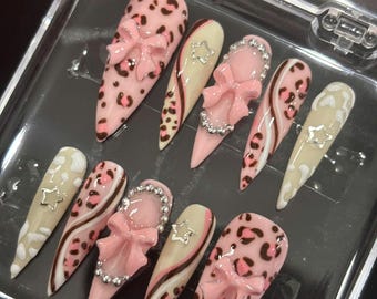 May include: A set of ten stiletto-shaped press-on nails in a clear plastic case. The designs feature a mix of light pink leopard print, cream-colored bases with brown swirls, 3D pink bows, silver star charms, and small silver metallic beads.