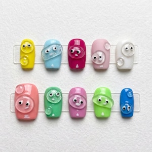 May include: A set of ten artificial nails in various colors, including yellow, blue, pink, and green. Each nail features a 3D design of a cartoon face with googly eyes and a clear, bubble-like accent. The nails are arranged on clear adhesive strips.