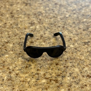 May include: A pair of miniature black plastic sunglasses with a classic aviator-style frame shape. The eyewear features dark, opaque lenses and curved temples designed for a small-scale figure, showcasing a solid black finish throughout the frame.