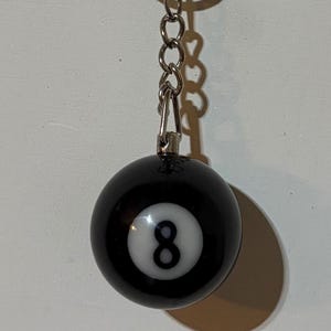 May include: A spherical black keychain featuring a white circular inlay with a black number eight printed in the center. The ball is attached to a silver-toned metal chain and a circular split ring for keys, creating a classic billiard ball design.