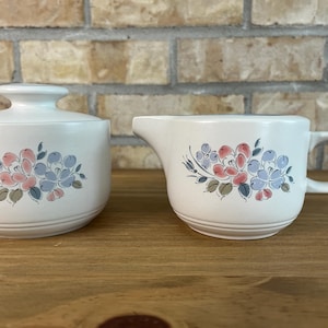 Peut inclure: Un sucrier en céramique blanche avec couvercle et un pot à lait assorti. Les deux pièces présentent un motif floral peint dans des tons de rose, bleu et vert, avec de subtiles rainures horizontales autour de la base de chaque récipient.