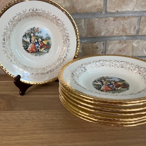 Peut inclure: Un ensemble de bols à soupe en céramique crème avec une illustration centrale représentant un couple dans un jardin, entouré d'une bordure en filigrane doré et d'un bord doré festonné. Un bol est présenté sur un support, les autres empilés.