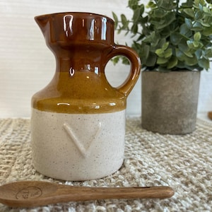 Peut inclure: Une cruche en céramique avec un dessus marron, un milieu beige et une base blanche mouchetée. Elle a une anse et un bec verseur. Une cuillère en bois avec le texte "TSP" est devant la cruche. Une petite plante en pot est en arrière-plan.