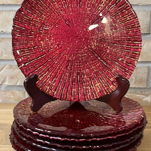 May include: A stack of circular glass plates featuring a deep red translucent base with a textured, radiating pattern highlighted by metallic gold accents. The plates have a scalloped, uneven edge and are displayed on a dark brown stand.