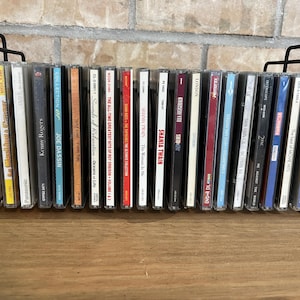 Könnte beinhalten: Ein schwarzer Metalldrahtständer mit einer Reihe verschiedener Musik-CDs in ihren Kunststoffhüllen. Der Ständer verfügt über ein einfaches rechteckiges Rahmendesign mit vertikalen Stützen an jedem Ende, um die Disc-Sammlung aufrecht zu halten.