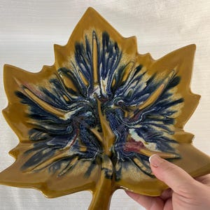Peut inclure: Un plat en céramique en forme de feuille avec une finition brillante. Le plat présente un extérieur marron et un centre avec des accents bleus, blancs et rouges. Le plat mesure environ 25 cm de long et 20 cm de large, avec une tige à la base.