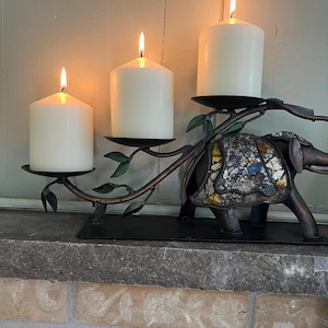 Peut inclure: Un bougeoir en métal en forme d'éléphant avec une couverture en mosaïque sur le dos. La structure présente trois branches incurvées s'étendant de l'éléphant, chacune surmontée d'une plaque circulaire supportant une bougie pilier blanche allumée.