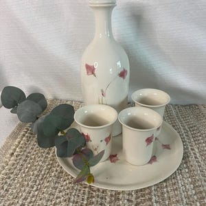 Peut inclure: Un service à saké en céramique blanche comprenant une bouteille haute et trois petites tasses, toutes ornées de motifs floraux roses délicats. L'ensemble comprend un plateau rond assorti. L'ensemble est posé sur une surface beige texturée.