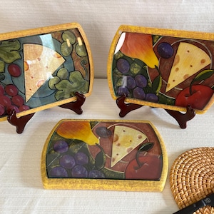 Peut inclure: Trois plateaux de service rectangulaires aux bordures texturées jaunes, chacun présentant un motif peint d'un morceau de fromage, de raisins et de fruits. Deux plateaux sont sur des supports marron, un est posé à plat près d'un dessous de plat.