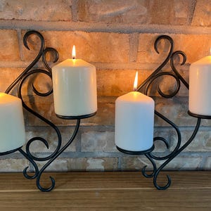 Peut inclure: Deux appliques murales en métal noir, chacune dotée d'un design incurvé en volutes et d'une plate-forme circulaire supportant une bougie pilier allumée. Les bougies blanc cassé émettent une flamme chaude et stable, créant une lueur vive.