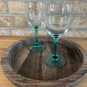 Peut inclure: Deux verres à vin en verre transparent dotés de pieds et de bases circulaires en vert émeraude translucide. Les verres présentent une forme de calice arrondie et lisse, posés sur un plateau circulaire en bois au grain sombre.