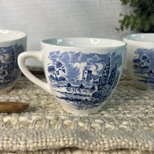 Peut inclure: Une tasse à thé en céramique blanche ornée d'un motif bleu de type transfert représentant un paysage avec un pont en pierre, un château et des arbres. La tasse possède une anse incurvée et une finition lisse, illustrant une scène rurale.