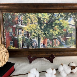 Peut inclure: Une peinture à l'aquarelle encadrée d'un bâtiment en briques rouges avec un balcon, un escalier et un vélo, entouré d'arbres verts luxuriants. Le cadre est de couleur marron foncé. Un panier en osier, un livre et des boules de coton sont au premier plan.