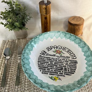 Peut inclure: Un bol à spaghetti en céramique avec un bord vert et blanc, avec du texte italien et des motifs floraux. Un moulin à poivre en bois, une salière et des couverts sont également présents, ainsi qu'une petite plante en pot.