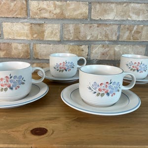 Peut inclure: Trois tasses à thé en céramique blanc cassé avec soucoupes assorties, ornées d'un motif floral peint à la main dans des tons de bleu doux, d'orange sourd et de vert. Les tasses présentent une forme arrondie avec une anse et un liseré bleu.