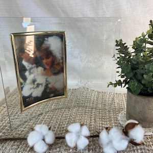 Pode incluir: Uma moldura de foto de vidro transparente com uma borda curva dourada, contendo uma foto de casamento. A moldura é apoiada por um suporte branco. Uma pequena planta em vaso e caules de algodão estão no fundo.