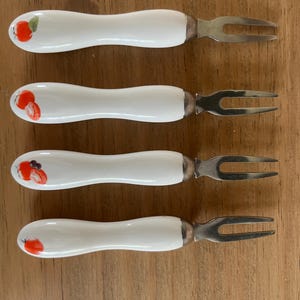 Peut inclure: Un ensemble de quatre fourchettes à apéritif avec des manches en céramique blanche, chacune ornée d'un petit motif de fruit peint. Les fourchettes possèdent des têtes en acier inoxydable à deux dents et un manche ergonomique incurvé.