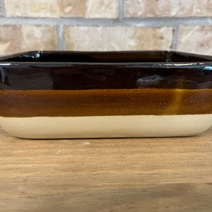 Peut inclure: Un plat de cuisson rectangulaire en céramique avec un vernis tricolore. Le bord supérieur est brun foncé, passant par une bande brun moyen au centre, et se terminant par une base de couleur crème claire sur la partie inférieure.