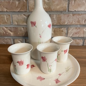 Peut inclure: Un ensemble de boissons en céramique composé d'une bouteille haute à col étroit, de deux gobelets assortis et d'un plateau de service rond. Chaque pièce est blanc cassé avec une finition émaillée craquelée et des motifs floraux roses peints.