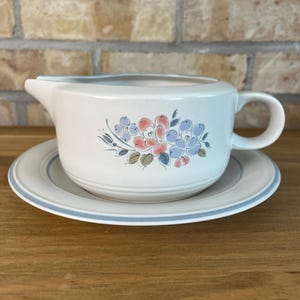 Peut inclure: Une saucière en céramique de couleur crème avec sa soucoupe assortie. La saucière présente un motif floral avec des fleurs roses et bleues et des feuilles vertes sur le côté. La soucoupe a une fine bande bleue sur le bord et la saucière a une anse.