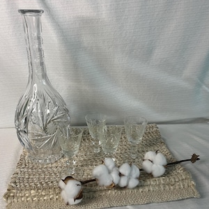 Peut inclure: Une carafe en verre transparent avec un long col et un motif en étoile sur le corps, accompagnée de quatre petits verres à pied assortis. Une branche décorative avec trois boules de coton blanc est placée devant le service en verre.