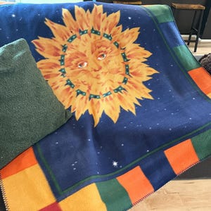 Peut inclure: Une couverture en polaire présentant un grand soleil jaune central avec un visage humain sur un fond bleu foncé parsemé de petites étoiles blanches. La bordure est composée de blocs rectangulaires alternant orange, jaune et vert.
