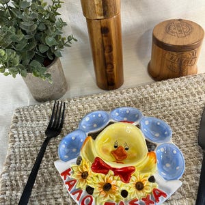 May include: A ceramic Easter plate with a yellow chick design, red bow tie, sunflowers, and blue egg-shaped dishes. The plate has the Greek text "Kαλό Πάσχα" in red. A wooden salt shaker and pepper grinder are visible in the background.