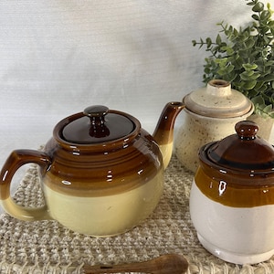 Op de afbeelding: Een keramische theeset in bruin en crème. De set bevat een theepot, een suikerpot en een melkkan. Een kleine houten lepel ligt voor de set. De items zijn gerangschikt op een geweven placemat.