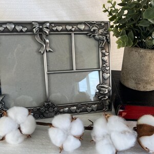 Peut inclure: Un cadre photo en métal argenté avec plusieurs ouvertures, décoré de cœurs, de nœuds et de petites figurines. Le cadre est à côté d'une petite plante en pot et de boules de coton.