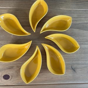 May include: A set of seven yellow ceramic dishes, each shaped like a teardrop or a leaf with a smooth, glossy finish. The dishes are arranged in a circular pattern, with the pointed ends facing outward, showcasing their uniform color and shape.