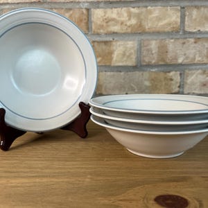 Peut inclure: Un ensemble de quatre bols en céramique blanc cassé avec une fine bande bleue simple entourant le bord intérieur. Un bol est présenté debout sur un support marron foncé, tandis que les trois autres sont empilés proprement à côté.