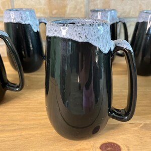 Peut inclure: Un ensemble de cinq tasses en céramique avec une finition noire brillante sur la partie inférieure et un émail gris clair texturé et moucheté sur le bord. Chaque tasse présente une forme cylindrique haute avec une anse latérale robuste.