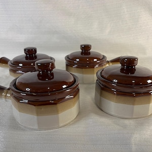 Peut inclure: Quatre bols à soupe en céramique marron et blanche avec couvercles. Chaque bol a une poignée et un couvercle marron foncé avec un petit bouton. Les bols ont une forme hexagonale et sont empilés sur une surface blanche.