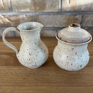 Peut inclure: Un pot à lait en céramique et un sucrier assorti avec couvercle, tous deux dotés d'un émail couleur crème avec des taches brunes. Le pot à lait possède une anse incurvée et un bec verseur, tandis que le sucrier inclut un couvercle amovible.