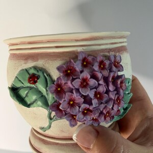 May include: A cream-coloured ceramic pot featuring a raised, three-dimensional cluster of purple hydrangea flowers with red centres. A green leaf with a small red ladybird is attached to the side of the pot, which has a rounded base and rim.