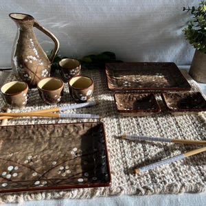 Peut inclure: Un ensemble de service à saké en céramique marron, comprenant une carafe assortie, quatre tasses et des plats de service. L'ensemble présente un motif floral avec des accents blancs. Des baguettes sont incluses, disposées sur un napperon tissé.