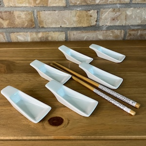 Peut inclure: Un ensemble de cinq repose-baguettes en céramique blanche avec une forme de bassin peu profond et allongé, accompagné d'une paire de baguettes en bois avec un motif à pois noirs et blancs sur les manches et des pointes marron clair.