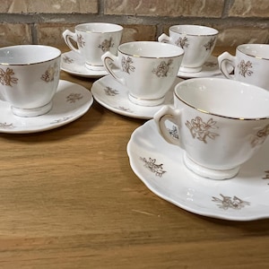 Peut inclure: Un ensemble de cinq tasses à thé en porcelaine blanche avec soucoupes assorties, ornées d'un motif floral doré délicat et d'un bord doré. Les tasses présentent une base sur pied et une anse incurvée, avec des soucoupes à bord festonné.