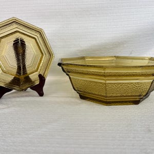 Peut inclure: Une assiette en verre ambré de forme octogonale avec un bord texturé, présentée sur un support sombre, accompagnée d'un bol octogonal en verre ambré assorti avec une surface extérieure texturée et une base intérieure lisse.