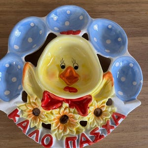 Könnte beinhalten: Ein Eierhalter aus Keramik mit einem zentralen gelben Küken mit roter Fliege, umgeben von sechs hellblauen Eierbechern mit weißen Punkten. Der Boden ist mit gelben Sonnenblumen und dem roten griechischen Text "ΚΑΛΟ ΠΑΣΧΑ" verziert.