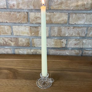 May include: A tall, slender, cream-coloured taper candle is lit, with a visible orange flame at the top. The candle is held upright in a clear, pressed glass candle holder that features a small circular handle and a decorative scalloped edge base.