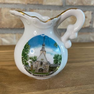 Peut inclure: Un petit pot à crème en porcelaine blanche avec un bord et une anse dorés. L'avant présente une illustration colorée d'une église en pierre avec un clocher, entourée d'arbres, avec le texte SANCTUAIRE NOTRE-DAME DU CAP inscrit en dessous.