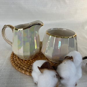 Peut inclure: Un ensemble de crémier et sucrier nacrés avec des bordures dorées. Le crémier a une anse et un bec verseur. Le sucrier est un petit récipient ouvert. Les deux sont sur un dessous de verre tressé, avec des boules de coton au premier plan.
